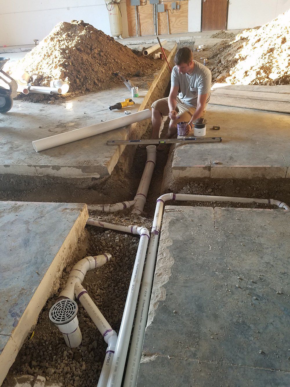 Plumber installing PVC pipes in a trench cut into a concrete floor, surrounded by gravel, tools, and dirt piles.