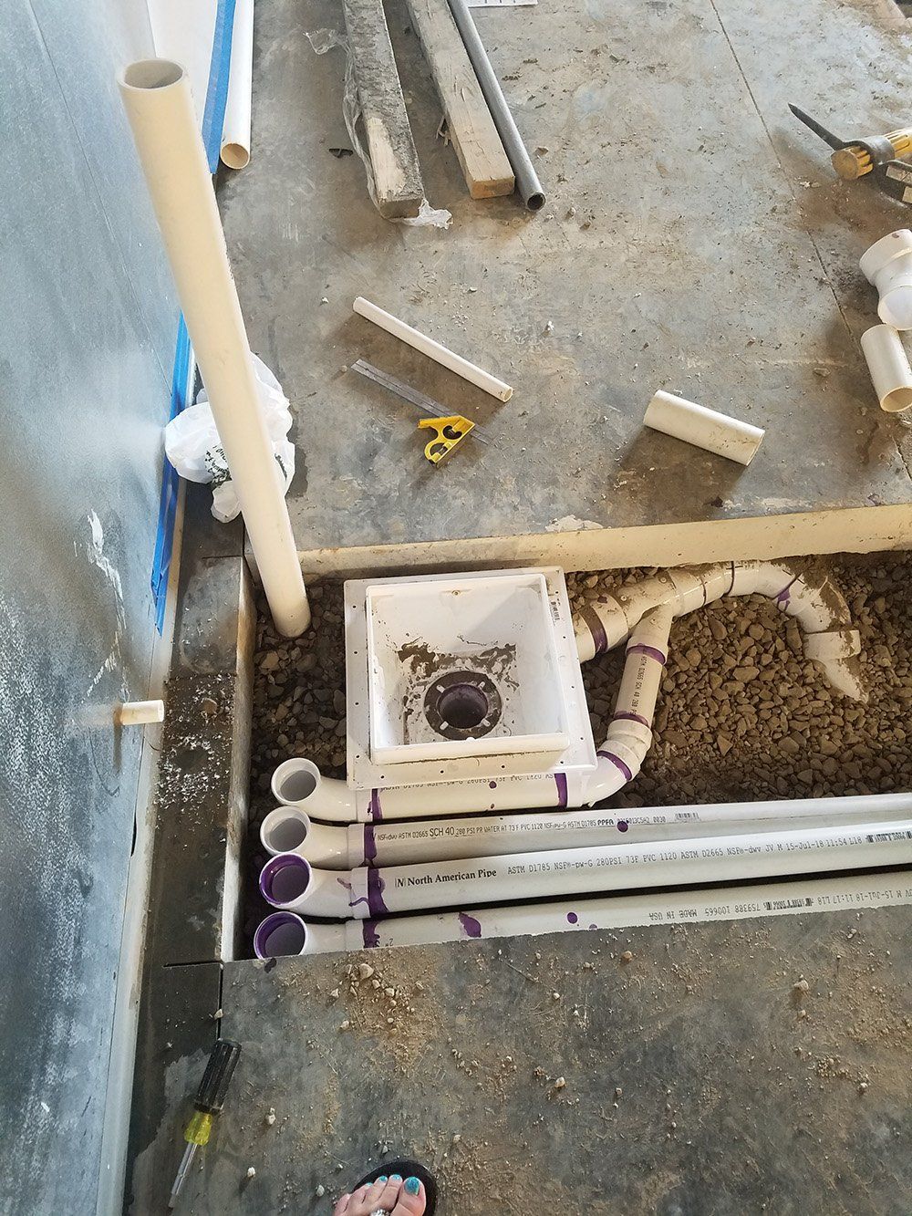 Construction site with exposed plumbing in a trench. White pipes and a drain box are installed in the floor, surrounded by gravel.