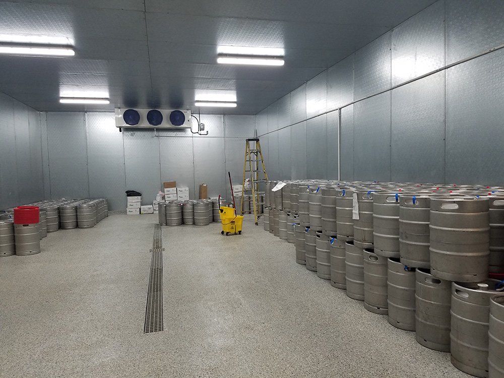 A large cold storage room filled with stacks of metal beer kegs, with overhead lighting and a small forklift.