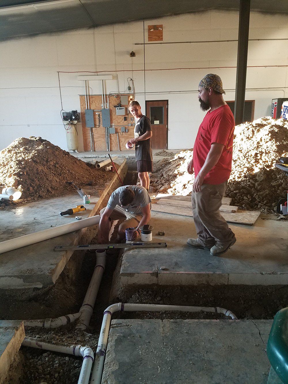 Three people working on pipes in a concrete floor. A person bends over the pipes, two watch. Construction site.