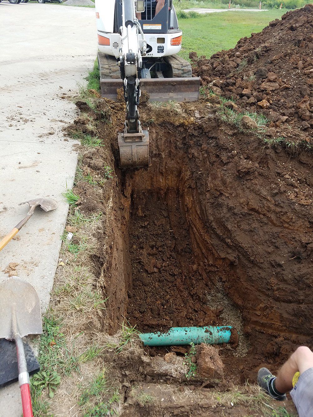 A small excavator digging a trench near a paved area, revealing a green pipe. Dirt piles up on the right.