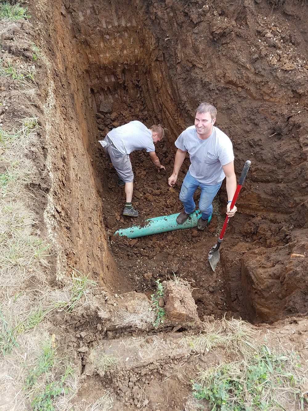 Two men in a deep dirt trench installing a green pipe. One is standing with a shovel, smiling; the other is bent over, working.