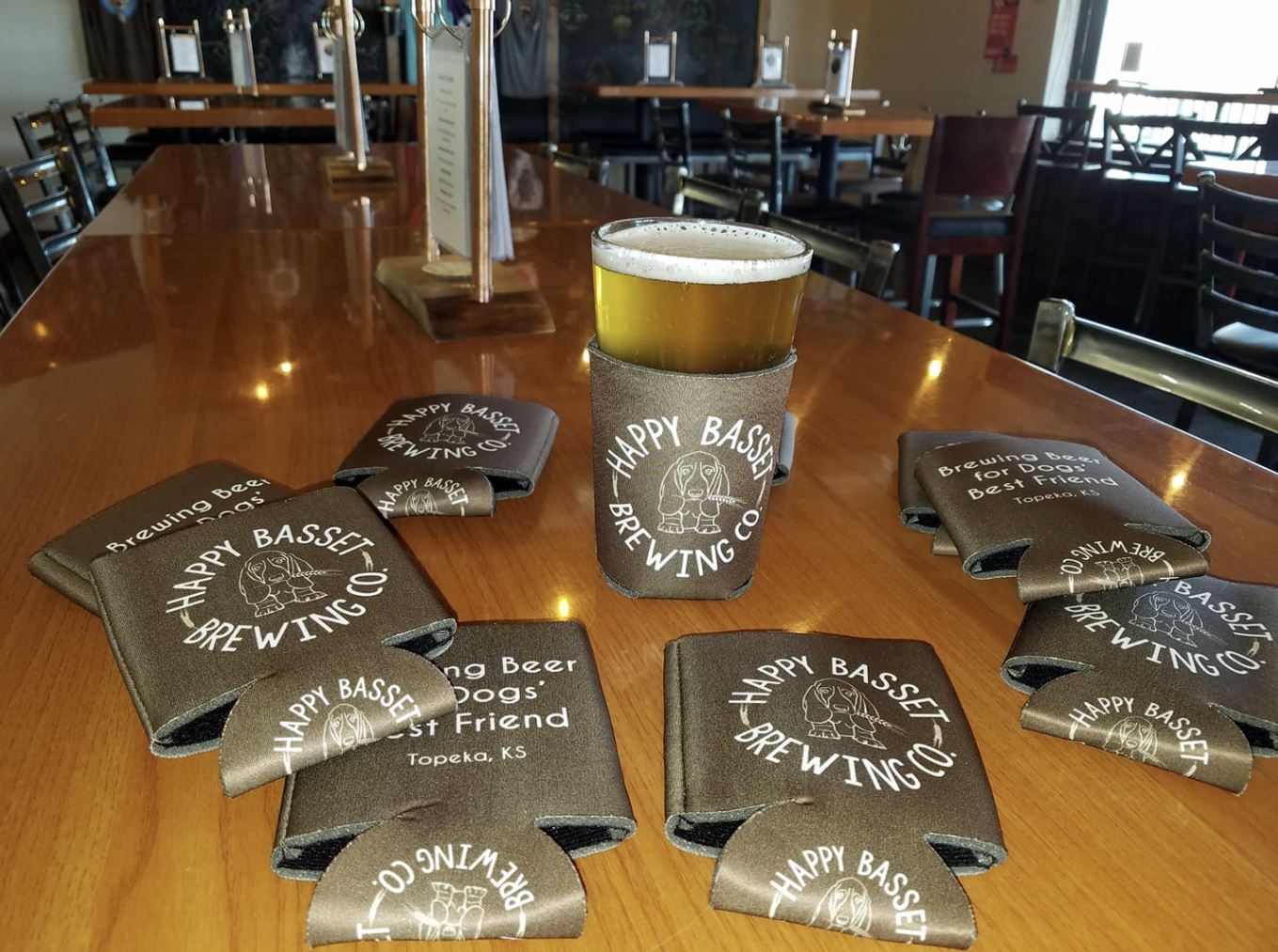 A beer glass in a Happy Bassett Brewing Co. koozie sits on a bar surrounded by more koozies.