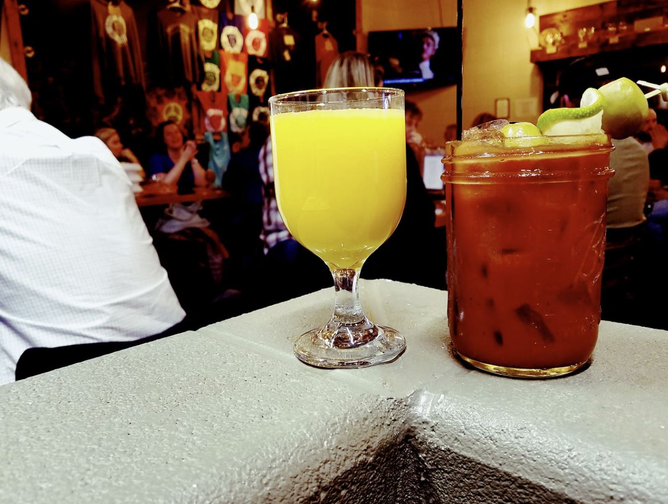 A yellow mimosa and red bloody mary sit on a counter at a bar. People are visible in the blurred background.