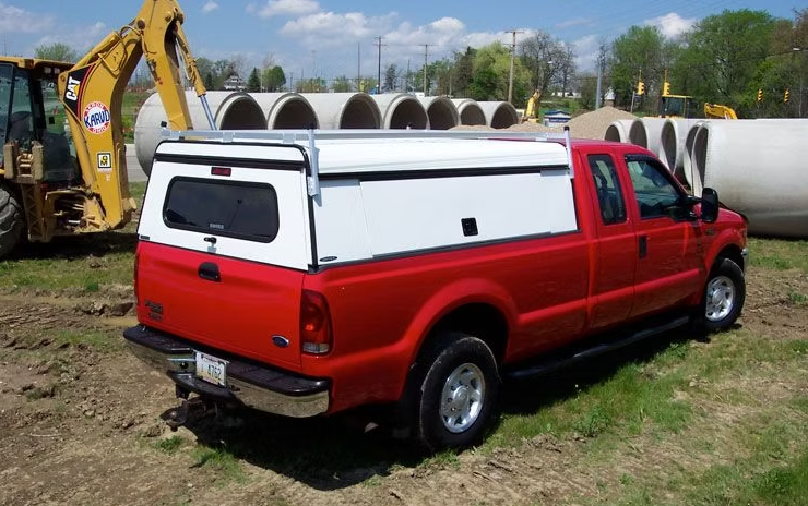 Red pickup truck with white bed cap, parked on grass near concrete pipes and construction equipment.