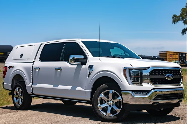 White Ford F-150 pickup truck with a white camper shell parked on gravel against a blue sky.