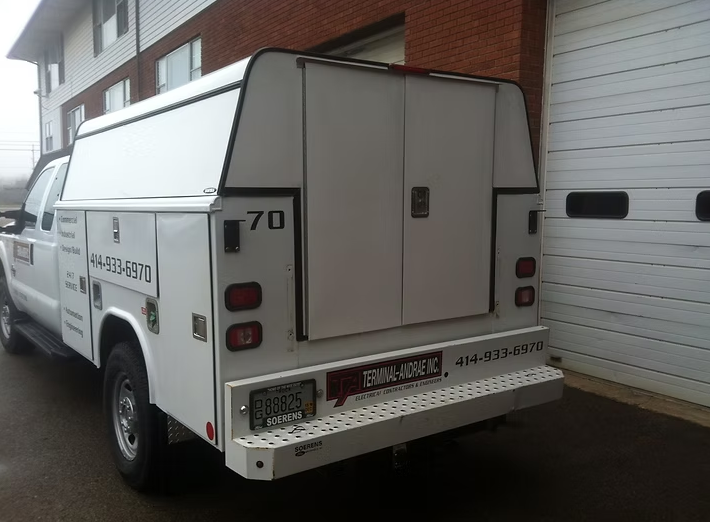 White service truck with a utility body, parked in front of a building with a roll-up door.