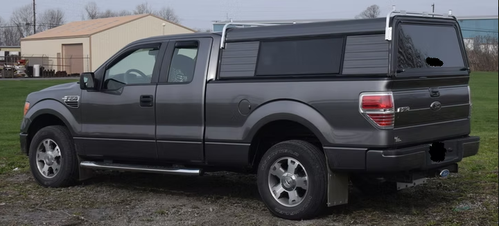 Gray Ford F-150 pickup truck with a black cap, parked on gravel in front of a tan building on a cloudy day.