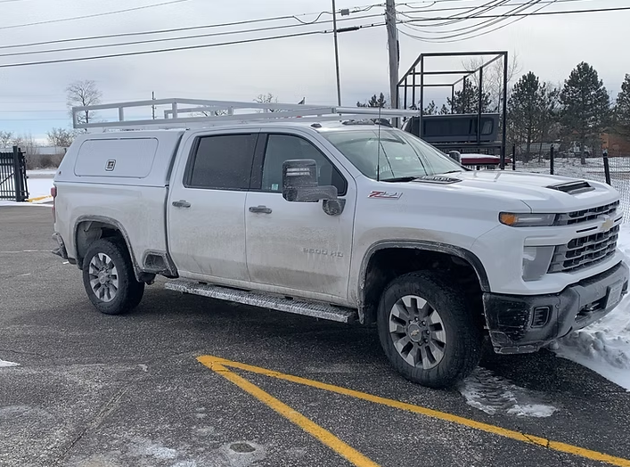 White Chevy pickup truck with cap and roof rack parked outdoors on a cold, snowy day.
