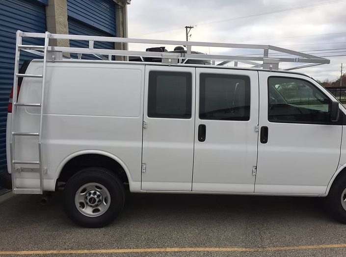 White work van with ladder and roof rack parked next to a blue building.