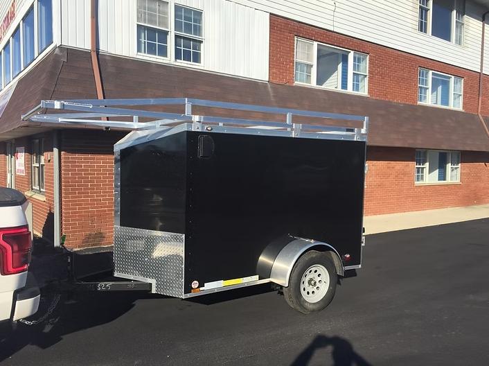 Black enclosed trailer with ladder rack hitched to a white truck parked near a brick building.