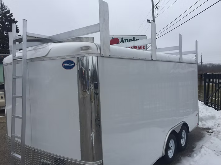 White enclosed cargo trailer with ladder and roof rack, parked outdoors.