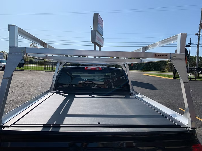 Truck bed with a silver ladder rack; black tonneau cover. Bright outdoor setting.