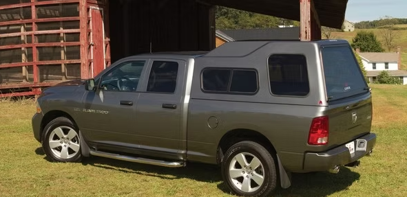 Gray Dodge Ram pickup truck with a cap parked next to a barn on grass.