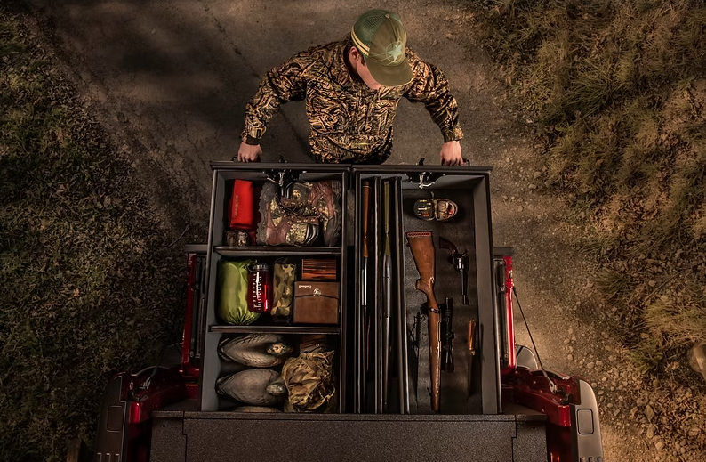 A person in camo gear looks into a truck bed storage system. The system holds hunting gear and a rifle.