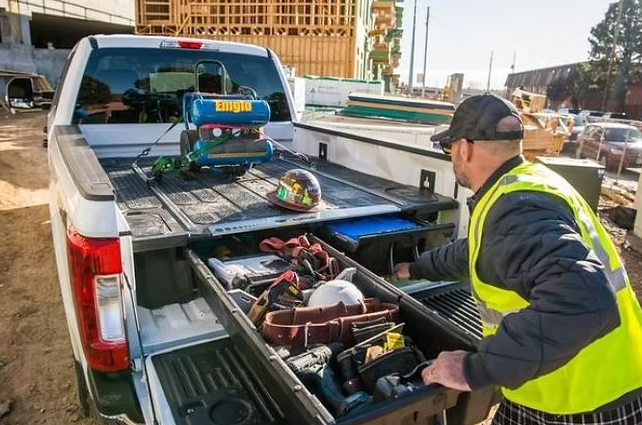 Construction worker using a truck bed drawer with tools, with a construction site in the background.