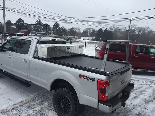 Silver Ford pickup truck with black bed cover, tool box, and ladder rack, parked in a snowy area.