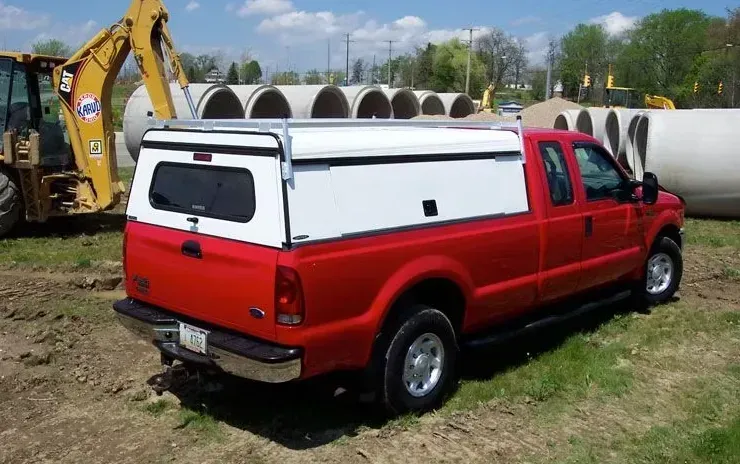 Red Ford pickup truck with white camper shell parked near concrete pipes and construction equipment.