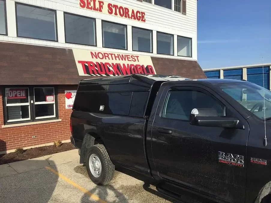 Black pickup truck with a camper shell parked in front of a Northwest Truckworld Self Storage building.