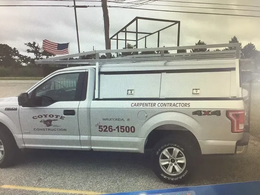 Silver work truck with ladder rack, storage compartments, and Coyote Contractors logo parked outdoors.