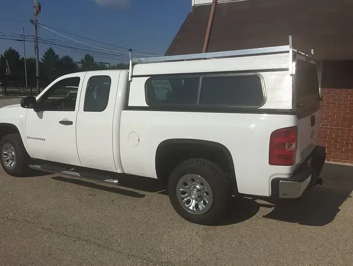 White pickup truck with a cap and roof rack parked on pavement next to a brick building.