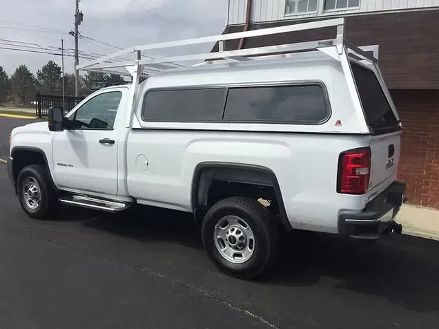 White pickup truck with a cap and roof rack parked on pavement next to a building.