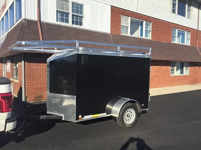 Black enclosed trailer with silver roof rack hitched to a white truck on asphalt.