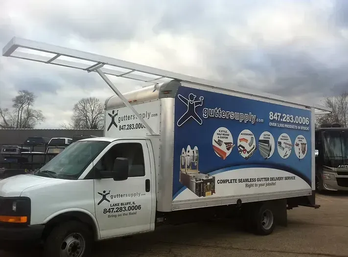 White box truck with blue and white graphics, extended metal rack, overcast sky. 