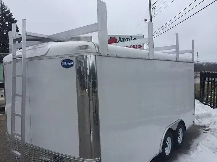 White cargo trailer with ladder and roof rack, parked near a storage facility.