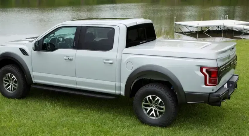 White Ford Raptor pickup truck with a tonneau cover parked on grass near water.