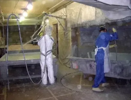 Workers in protective suits spray coating in a workshop, working near large vats under overhead lights.