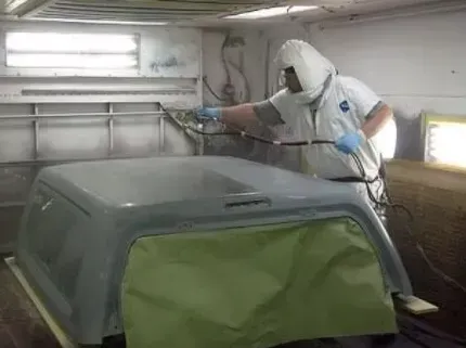 Person in protective suit spray painting a vehicle component in a spray booth.