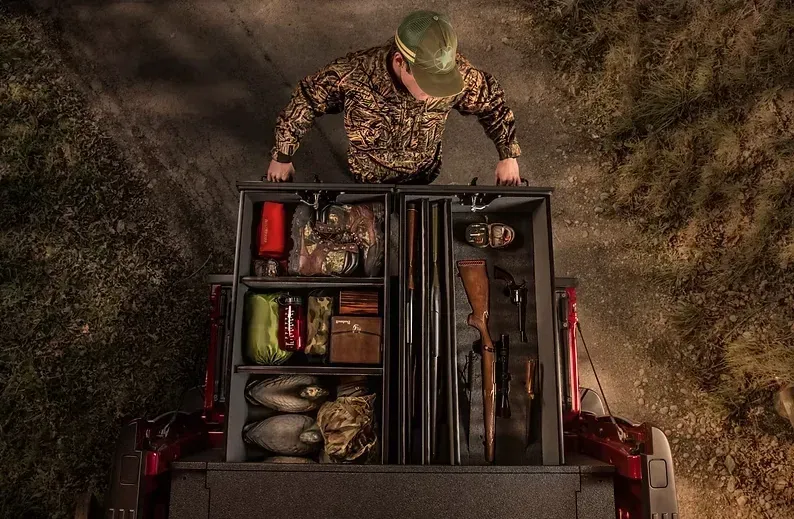 Person in camo gear opens a truck bed organizer with hunting gear and firearms.