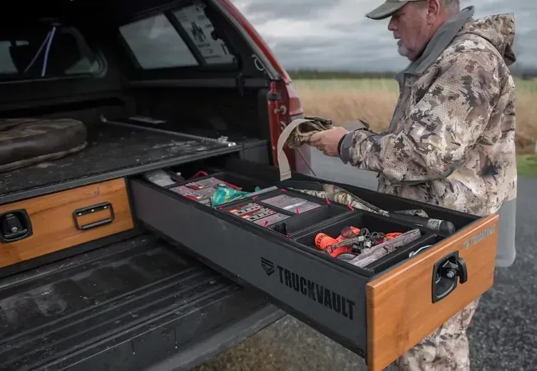 Man in camo opens truck drawer, filled with tools, next to open truck bed and field.