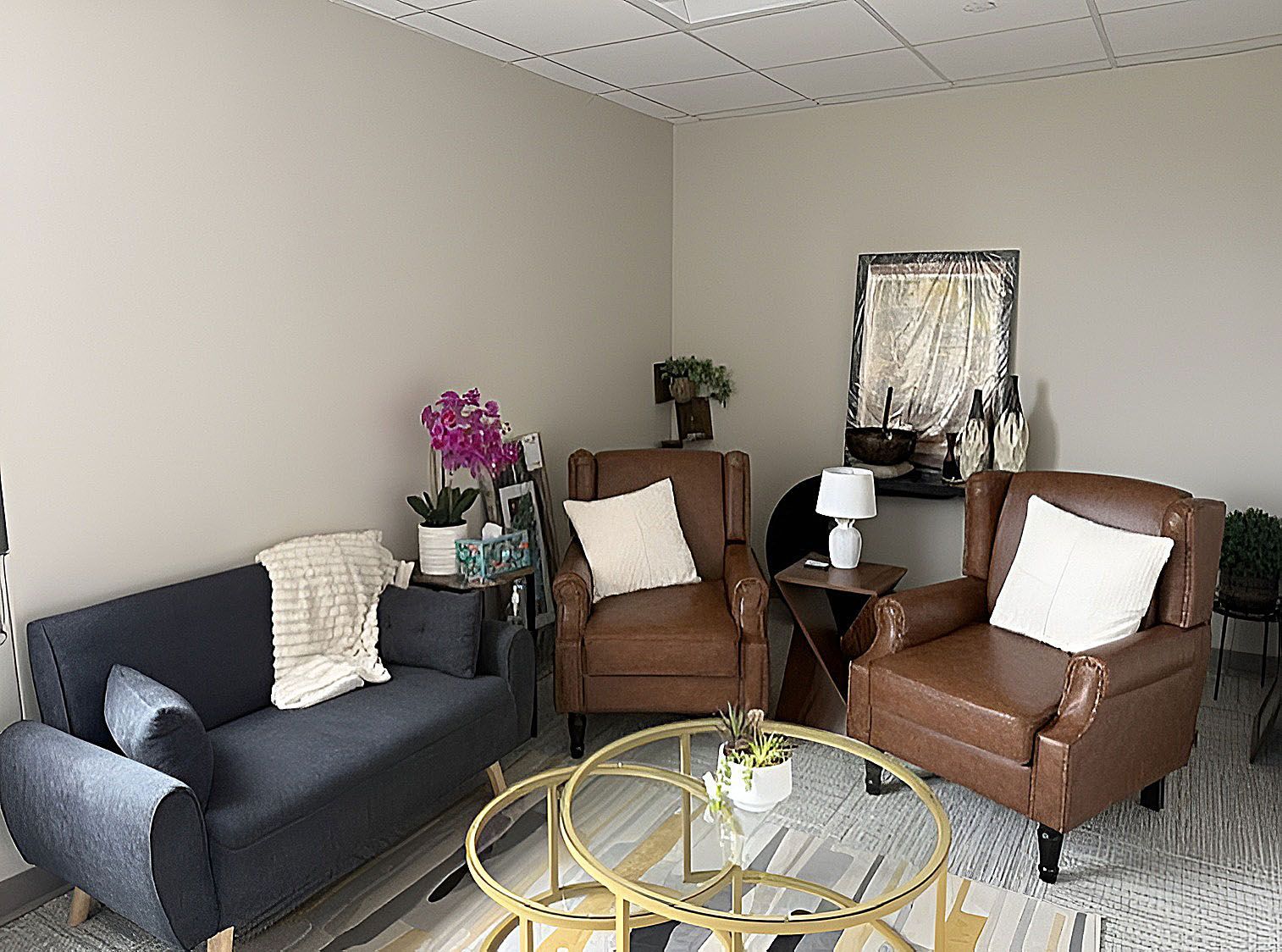 Cozy living room with gray sofa, two brown chairs, glass coffee table, and decorative plants.