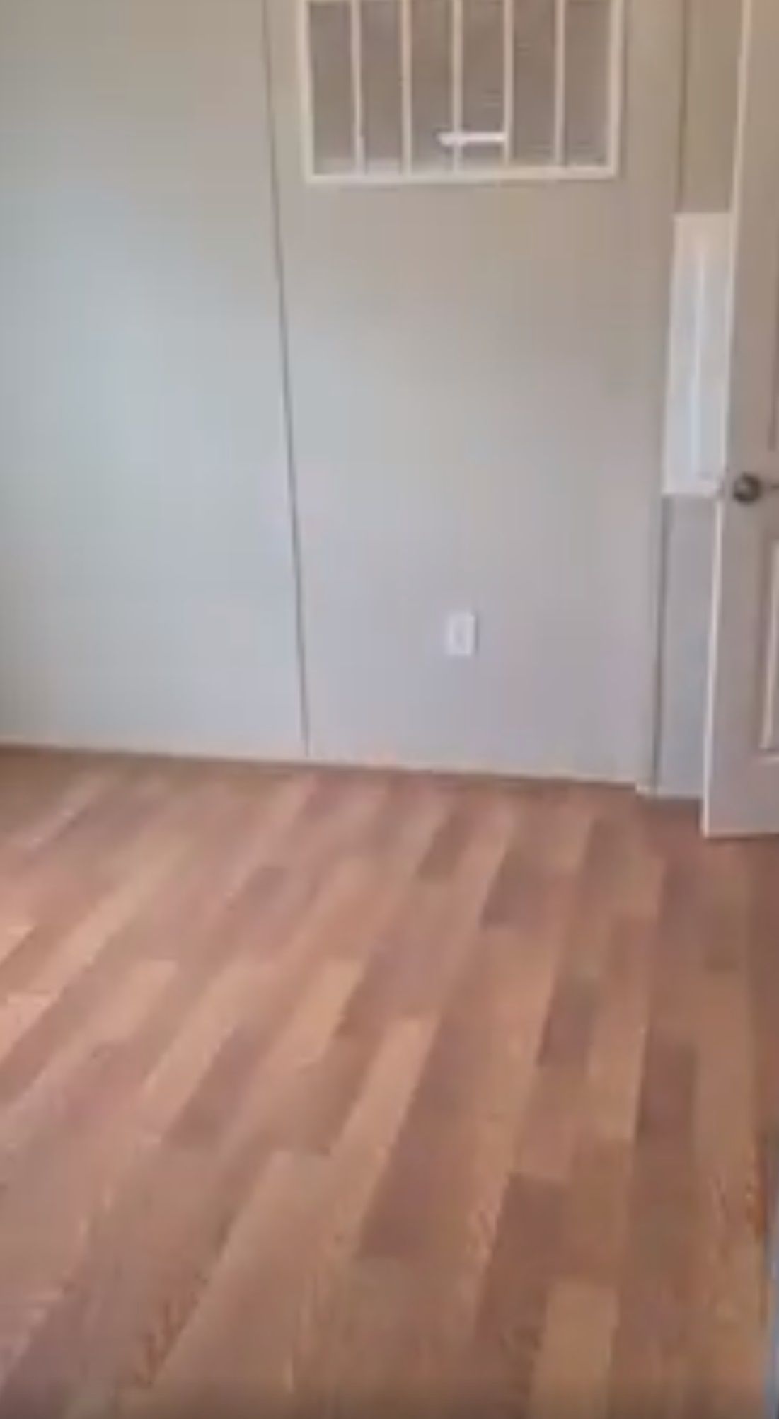 Empty room with wood-look flooring, light gray walls, a small vent, and a closed white door.