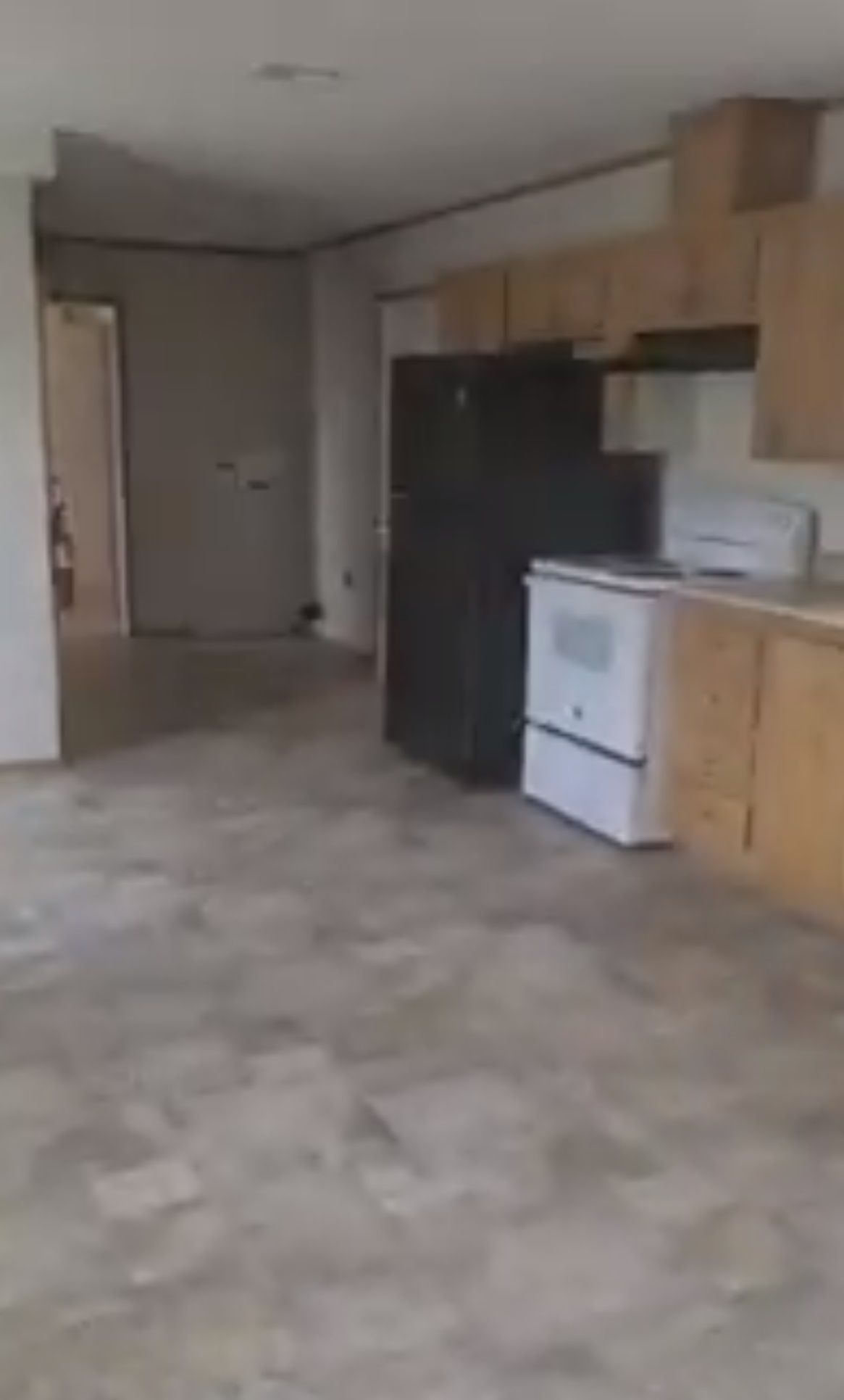 An empty kitchen and living area with appliances: stove, refrigerator, cabinets. Gray flooring, neutral walls.