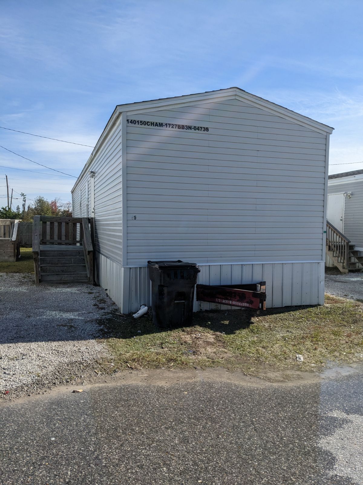 Gray mobile home exterior with steps, trash can, and trailer on gravel and grass.