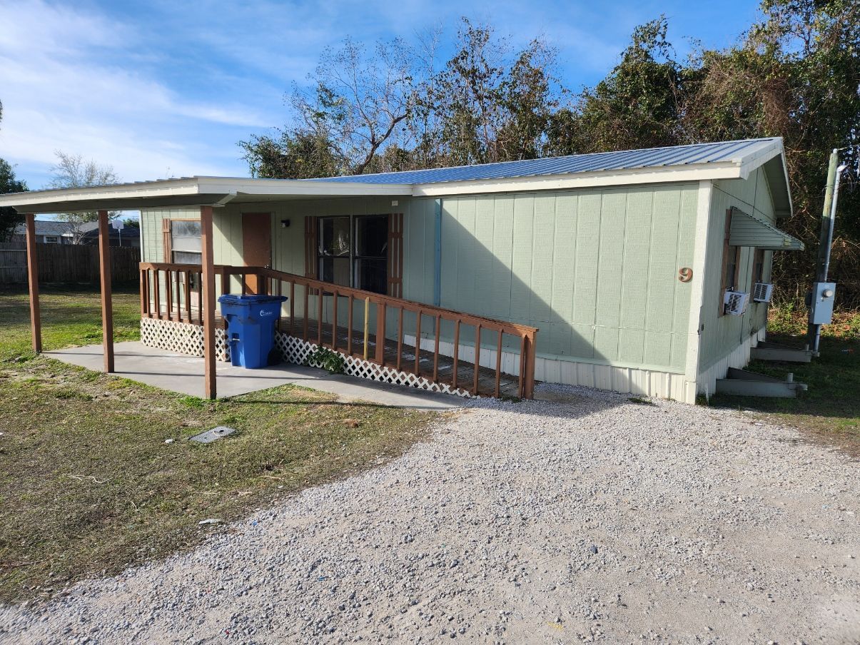 Green house with a ramp and covered porch. Gravel driveway. Blue sky.