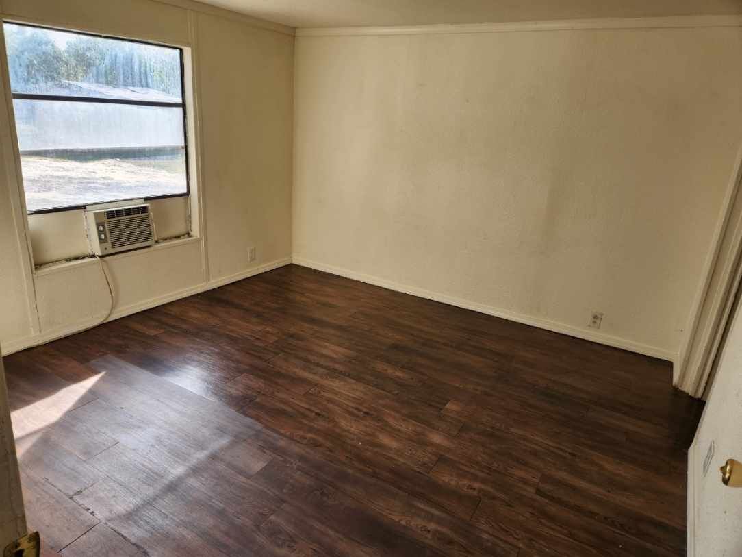 Empty room with dark wood flooring, beige walls, and window with air conditioner unit.