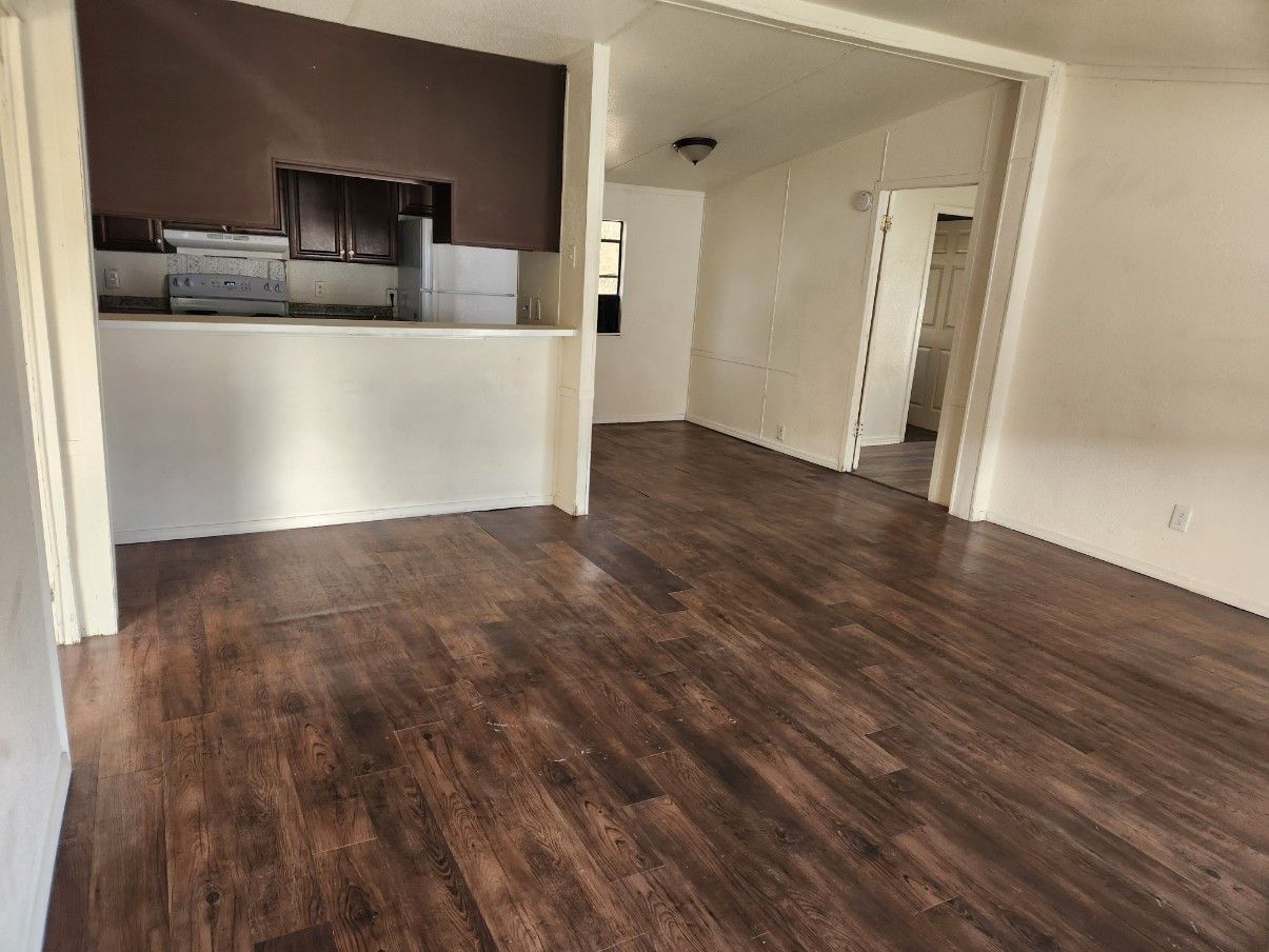Empty room with dark wood-look floor, kitchen in the background, doorways to other rooms.