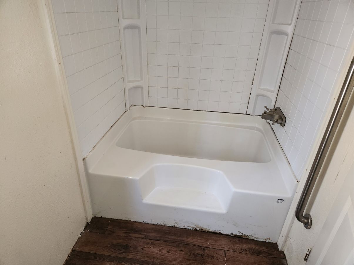White bathtub with step, tile surround, and grab bar, installed in a small bathroom with wooden flooring.