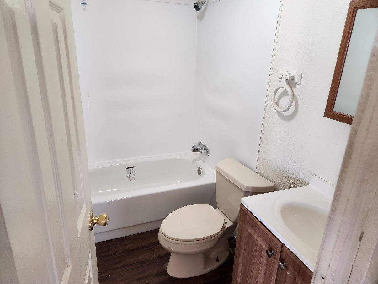 Small bathroom with white tub, toilet, and vanity. Brown cabinets, dark flooring, and a white door.