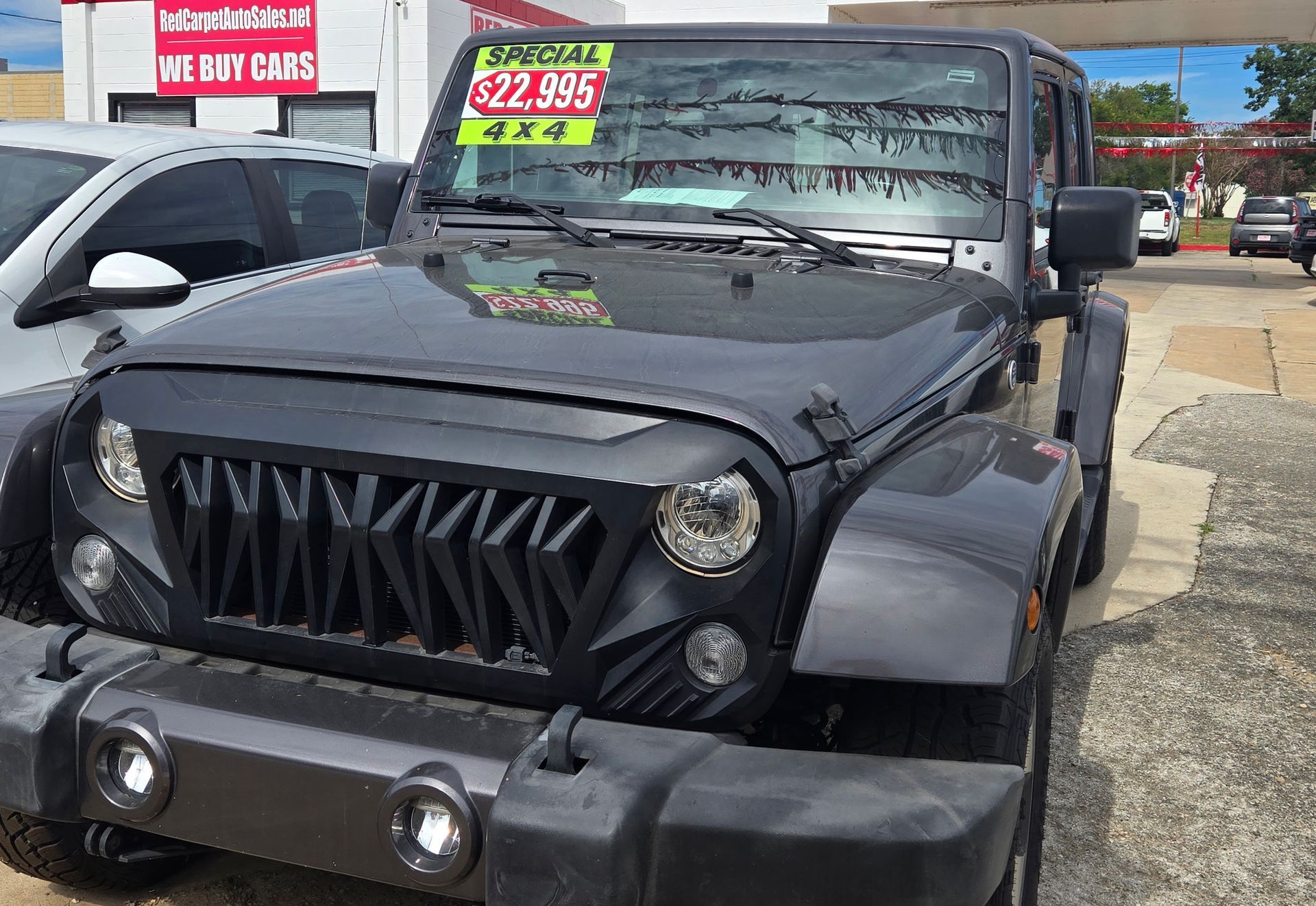 Dark gray Jeep Wrangler with a custom grill displayed on a car lot under a sunny sky. The price tag reads $32,995.
