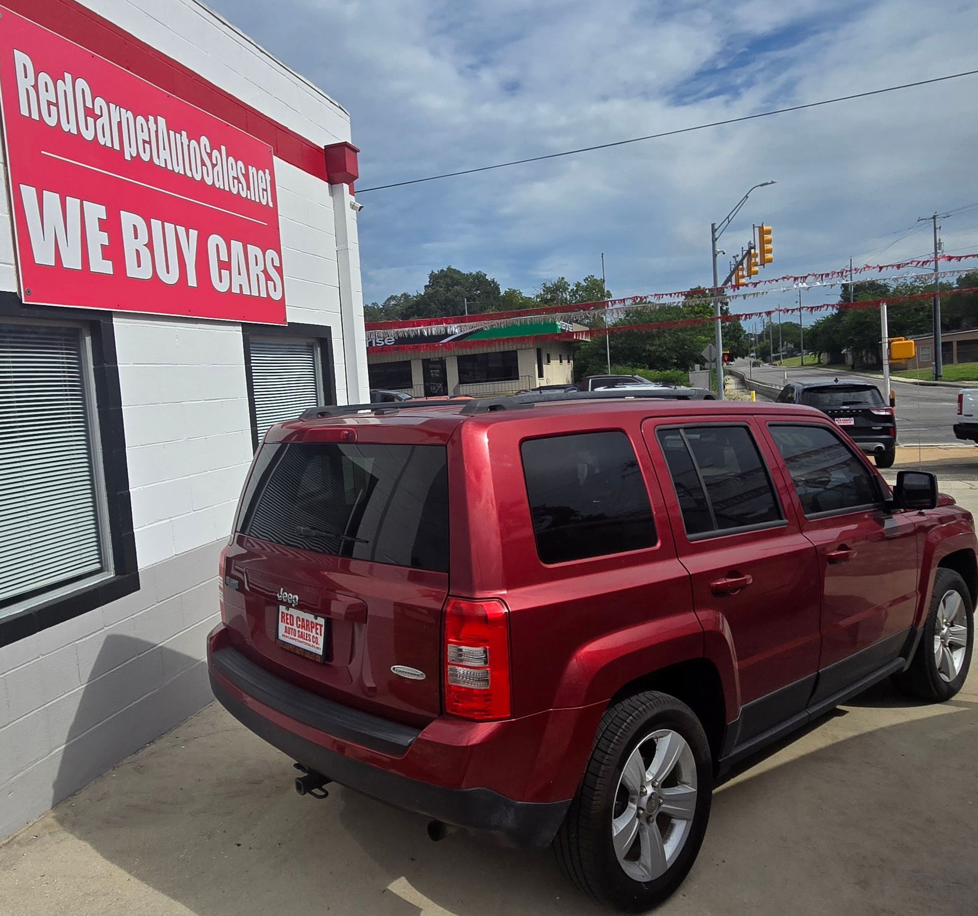 Red Jeep Patriot parked outside a car buying business with a red and white sign that says