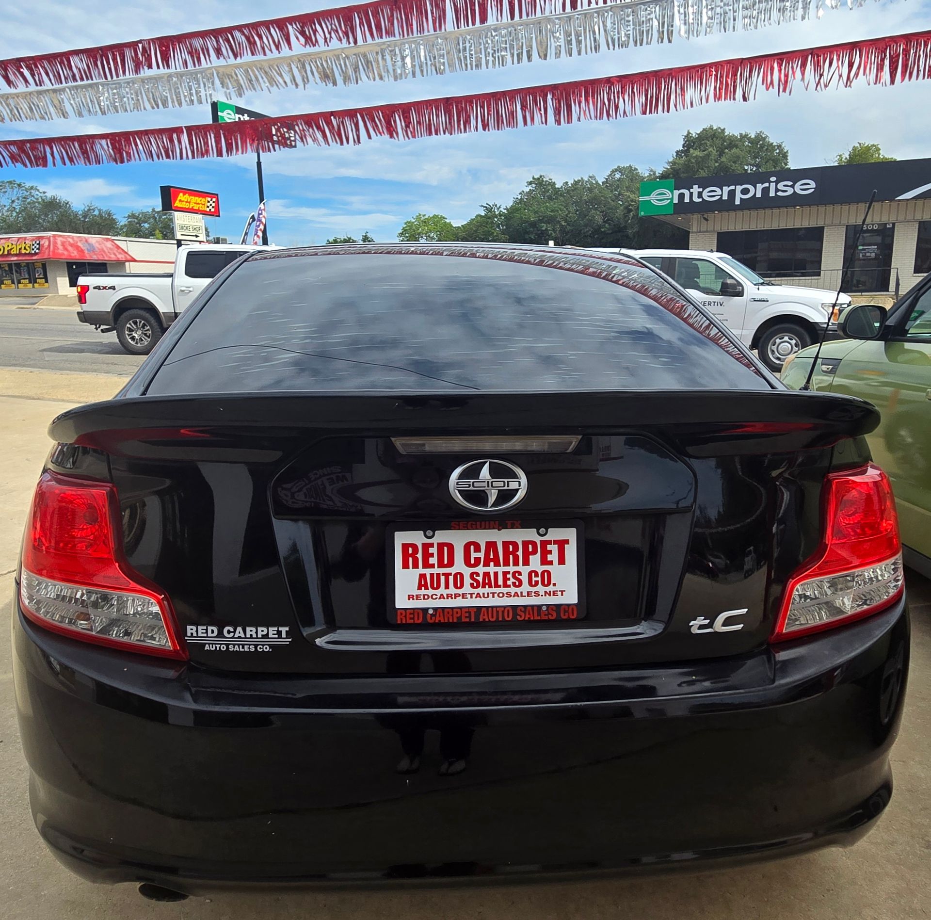 Black Scion TC rear view with Red Carpet Auto Sales plate, under red and white banners, parked outside a car dealership.