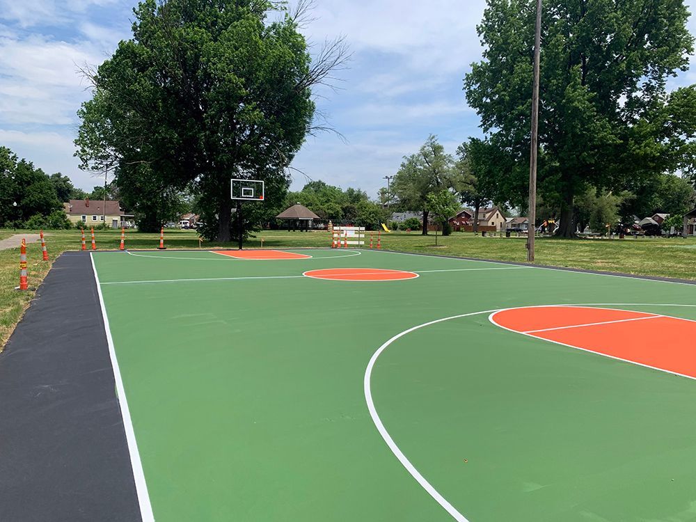A green and orange basketball court in a park with trees in the background.
