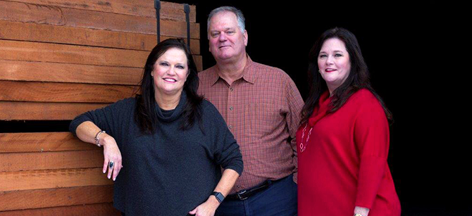 A man and two women are standing next to each other in front of a wooden wall.