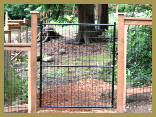 Black metal gate between wooden fence posts, leading to a path in a wooded area.