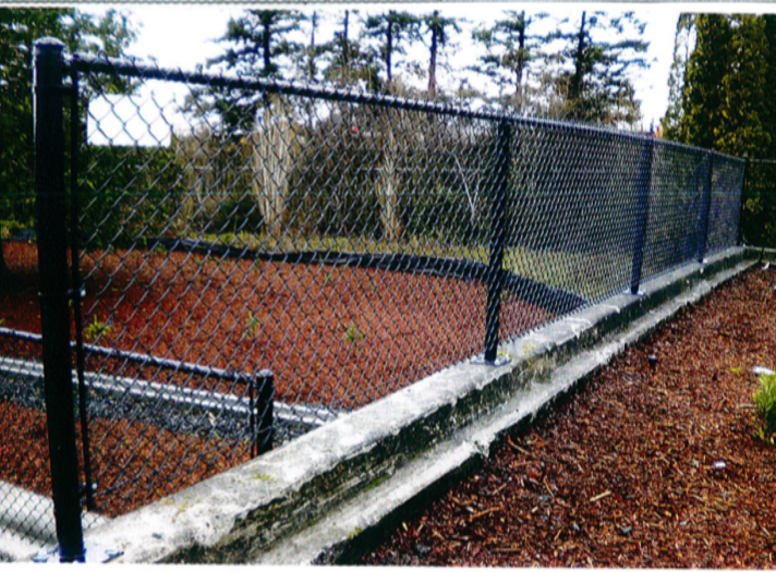 Black chain-link fence atop a concrete retaining wall, brown mulch in foreground, trees and bushes in the background.